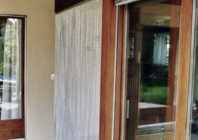Le rideau anti-mouches krismar monté devant une porte de terrasse. Les chaînes en aluminium du rideau se complètent optiquement très bien avec la construction en bois de la maison montrée.